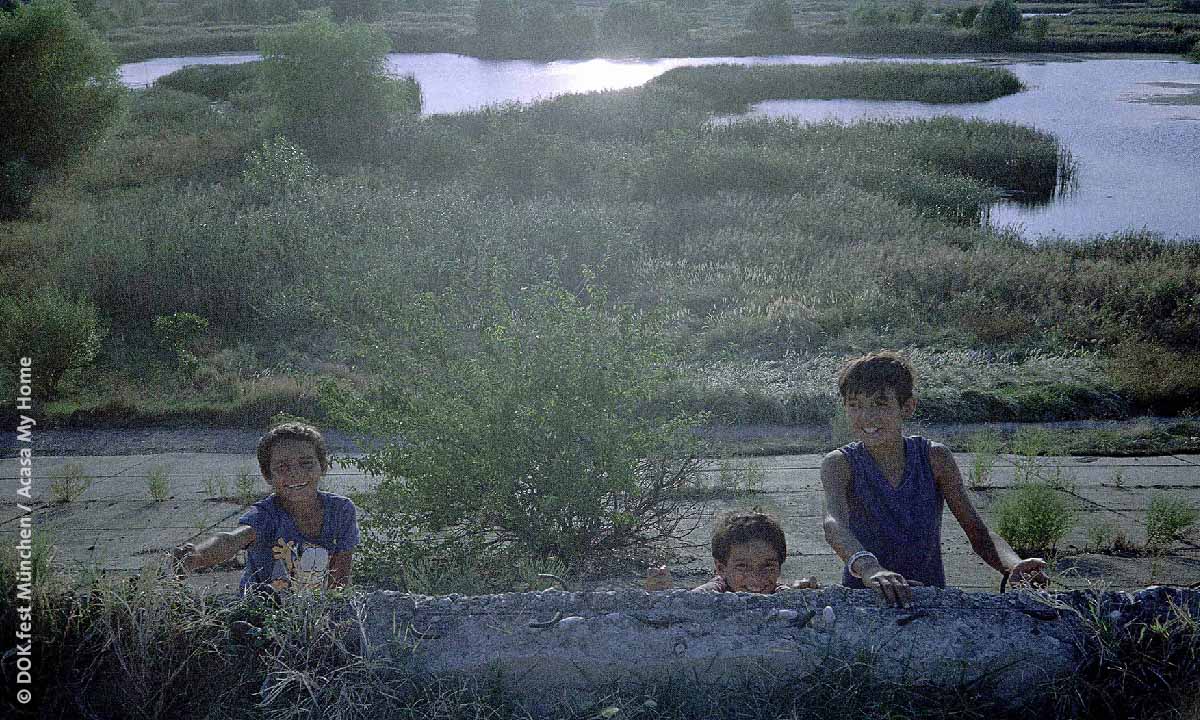 Filmstill aus „Acasa, my Home“: Drei Jungs hinter einem Mauerrest vor einer idyllischen Seenlandschaft
