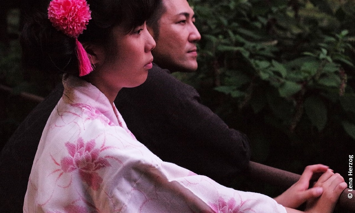 Filmstill aus "Family Romance, LLC": Bild eines japanischen Paares, die gemeinsam von einem Geländer aus in die Ferne blicken (© Lena Herzog)