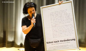 Schrei nach Veränderung bei der AG DOK: Bild einer Frau die ein großes Plakat hält. Sie hat ein Mikrofon in der Hand. (© AG DOK Südwest)