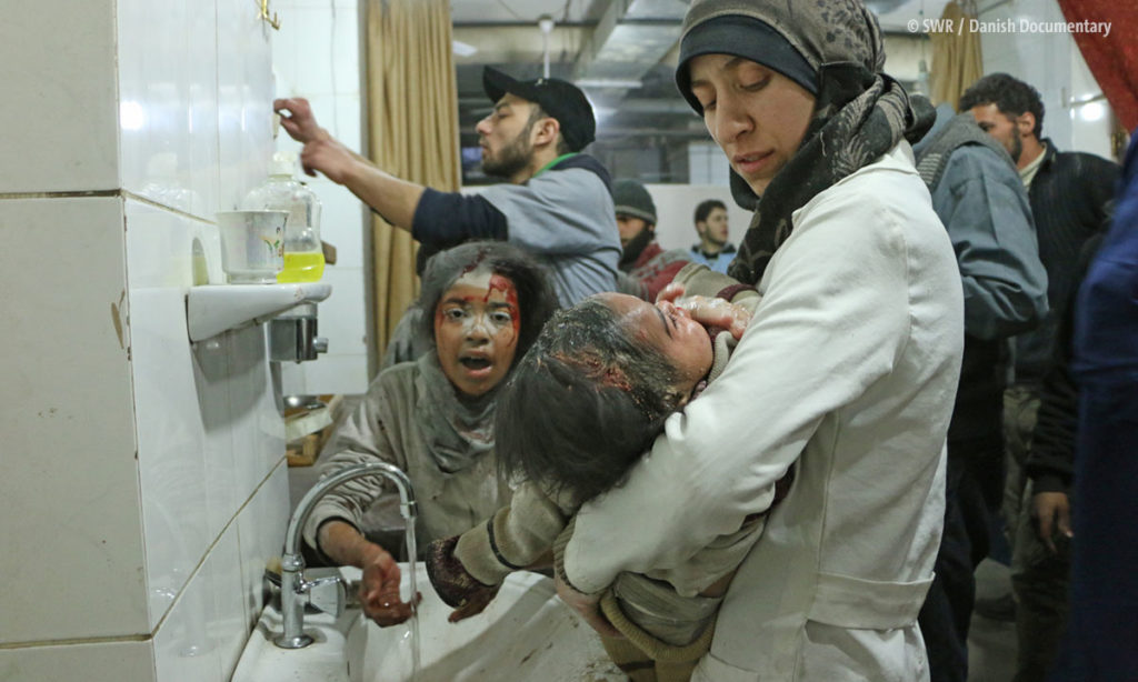 Filmszene aus "The Cave": Kriegsverletzte in einem Krankenhaus, Frau mit Kopftuch hält blutiges Kind in ihren Armen (© SWR / Danish Documentary)