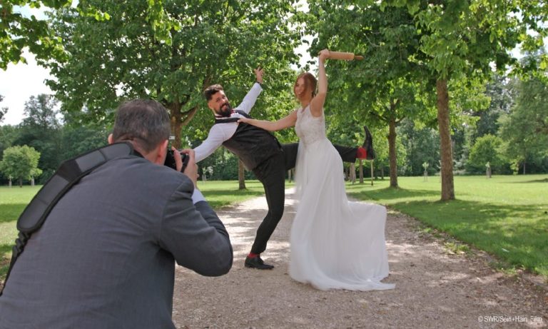 Filmstill aus "Und Küsschen": Foto eines fröhlichen Hochzeitspaares im Freien beim Fototermin