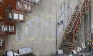 Loveparade – Die Verhandlung: Die Gedenkstätte für die Toten und Verletzten der Loveparade 2010 in Duisburg. Bild einer Wand mit Treppe auf der Kerzen und Graffiti zu sehen sind.